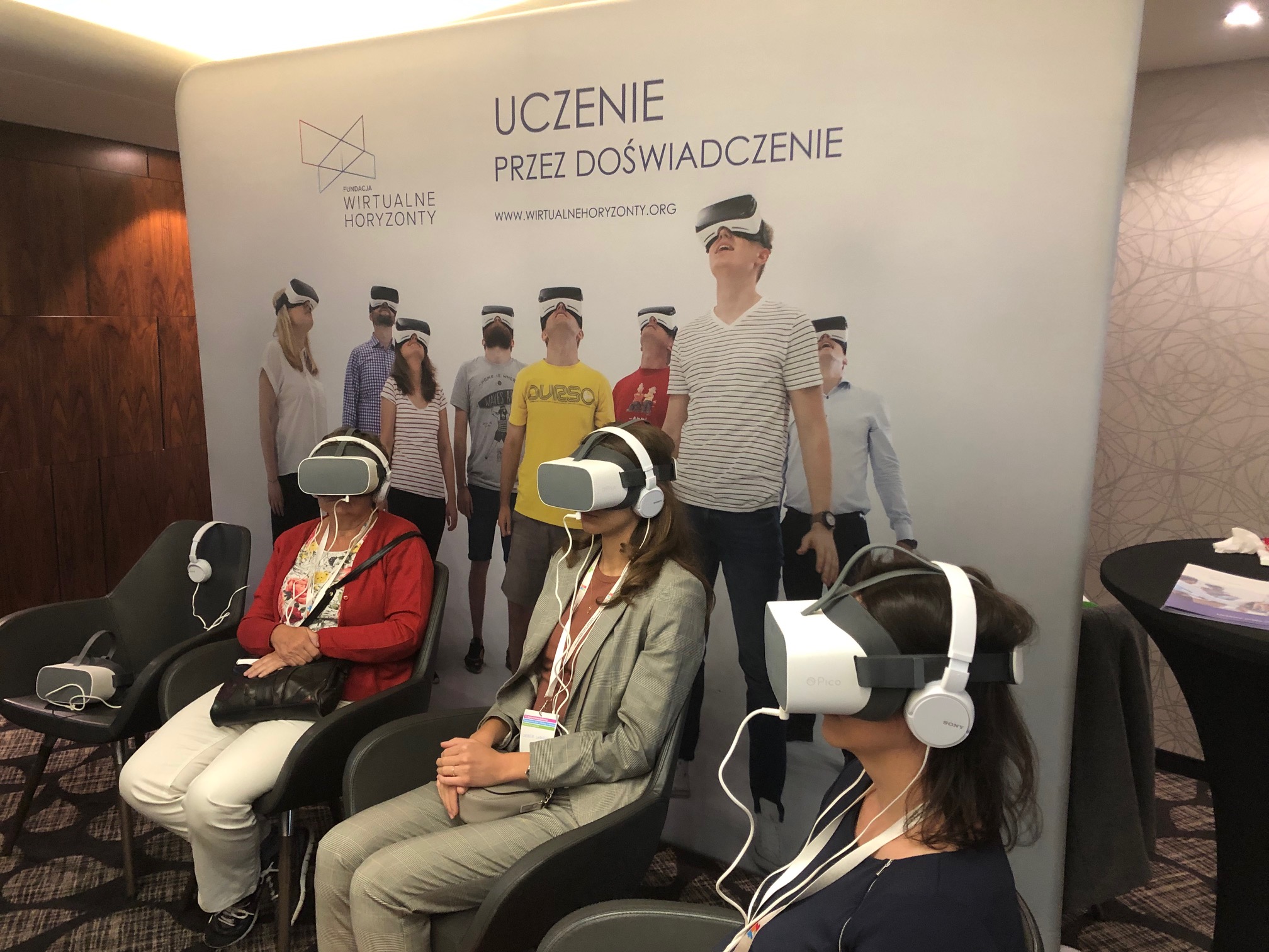 Trzy osoby z okularami VR i słuchawkami na uszach. W tle plakat: uczenie przez doświadczenie.