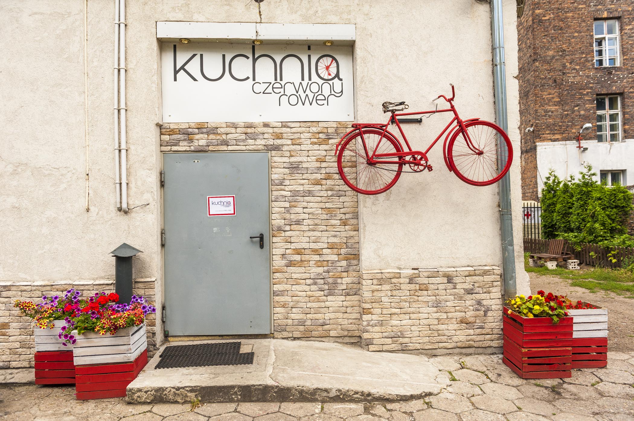 Zdjęcie przedstawia drzwi nad nimi napis Kuchnia Czerwony Rower. Na ścianie budynku wisi przyczepiony jako ozdoba rower.