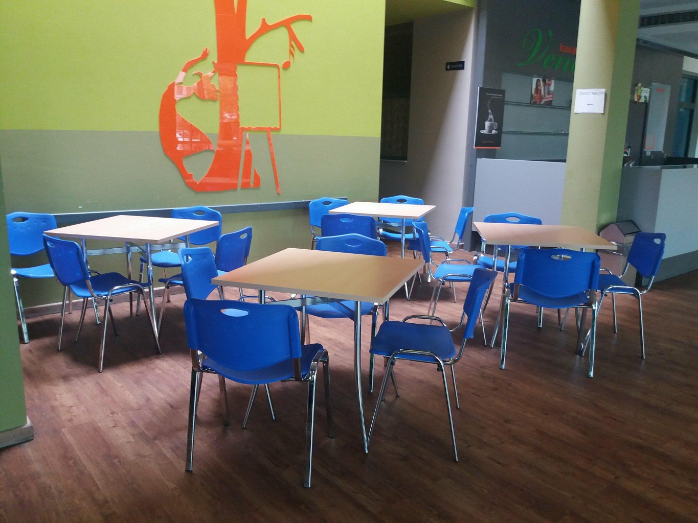 przestrzeń z kolorowymi ścianami, w której stoją cztery czteroosobowe stoły, a przy każdym krzesła.