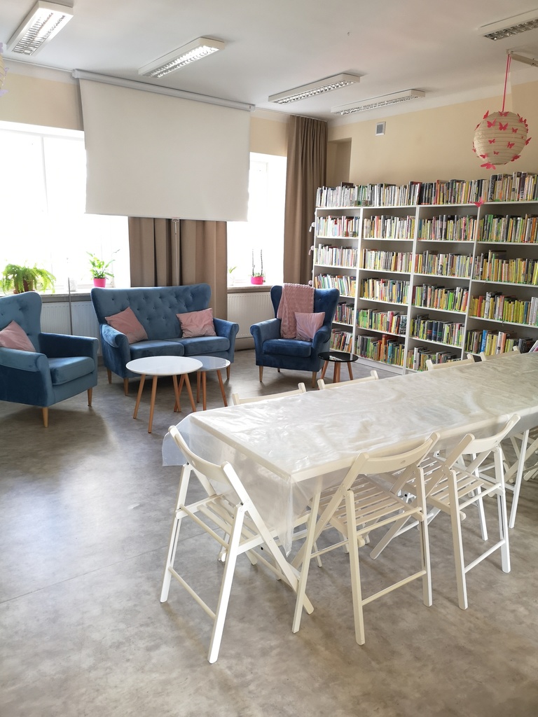 Pomieszczenie z kanapami, fotelami, podłużnym stołem i składanymi krzesłami. Przy ścianie regały z książkami.