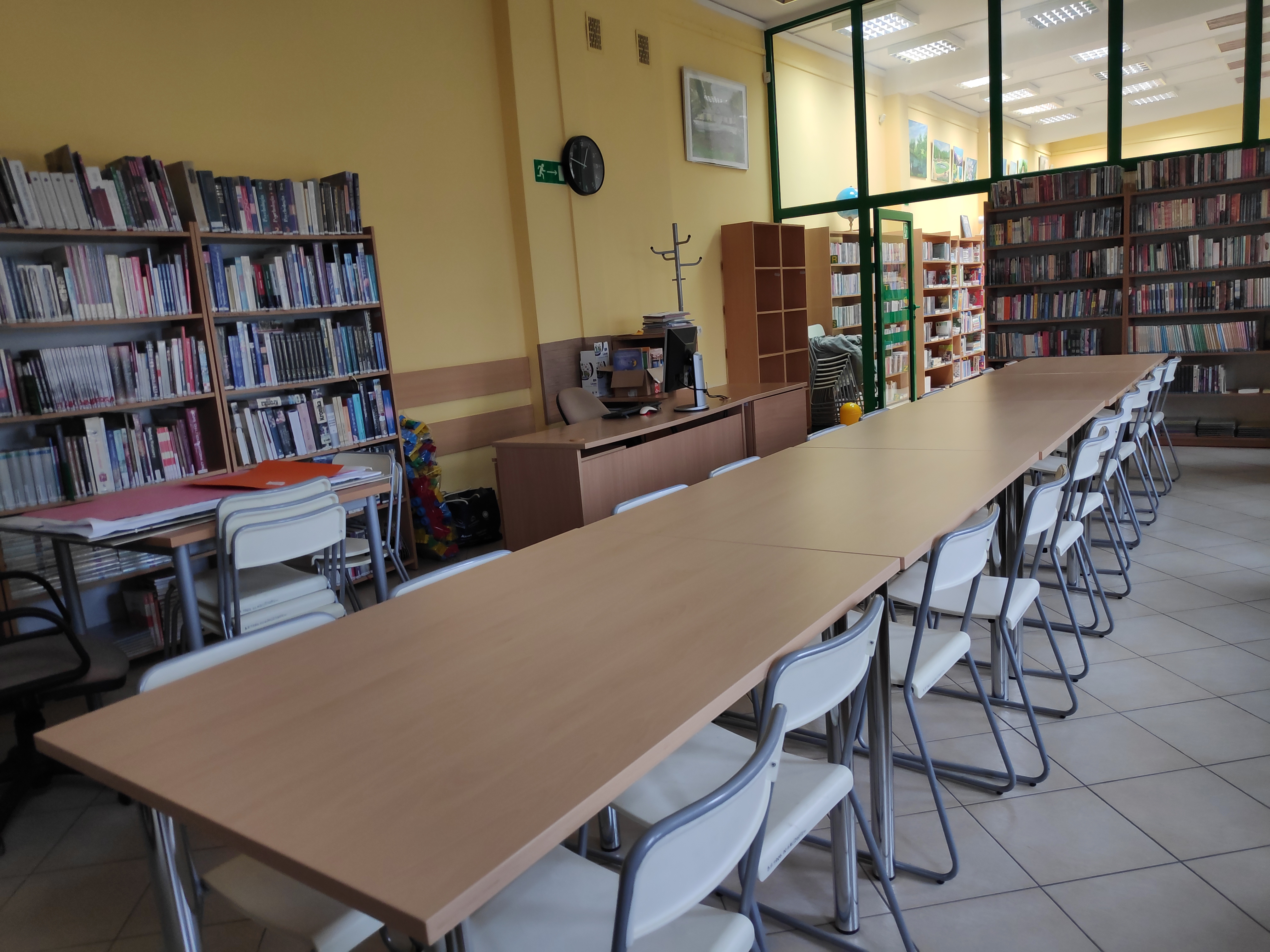 Długi stół z krzesełkami, ustawiony w sali. Pod ścianami regały z książkami.