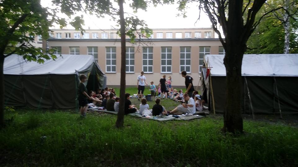 Zdjęcie pola biwakowego znajdującego się przy dużym budynku. Na polu namioty wojskowe i osoby biwakujące.