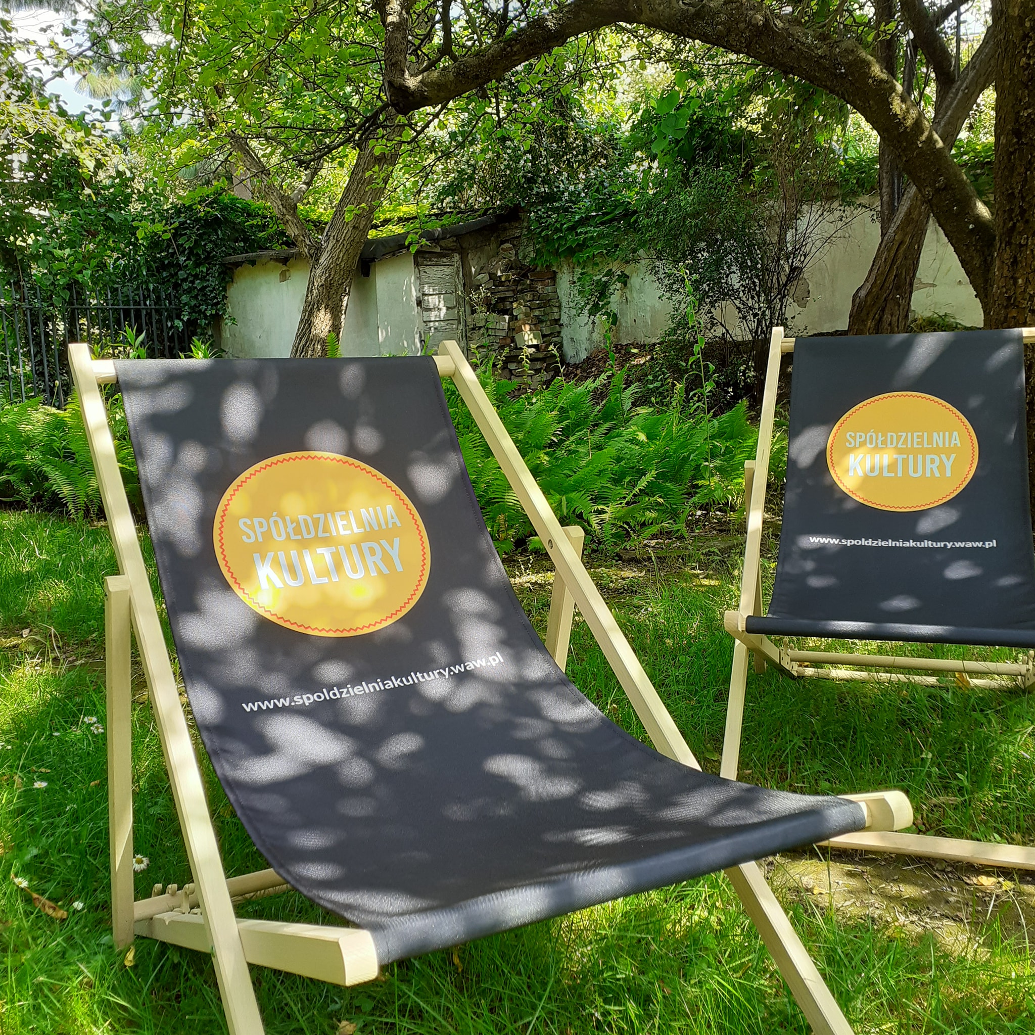 Dwa czarne leżaki z nadrukowanym pomarańczowym kołem i napisem Spółdzielnia kultury w miejscu, w którym można się oprzeć plecami. Leżaki ustawione są na trawie, w słońcu, w pobliżu drzew. 