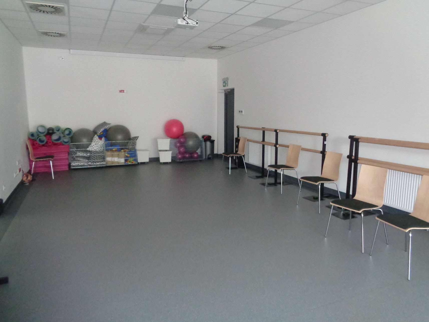 Podłużna sala ze sprzętem do ćwiczeń (np. gumowe piłki) znajdującym się pod ścianą. W sali znajduje się też kilka krzeseł.