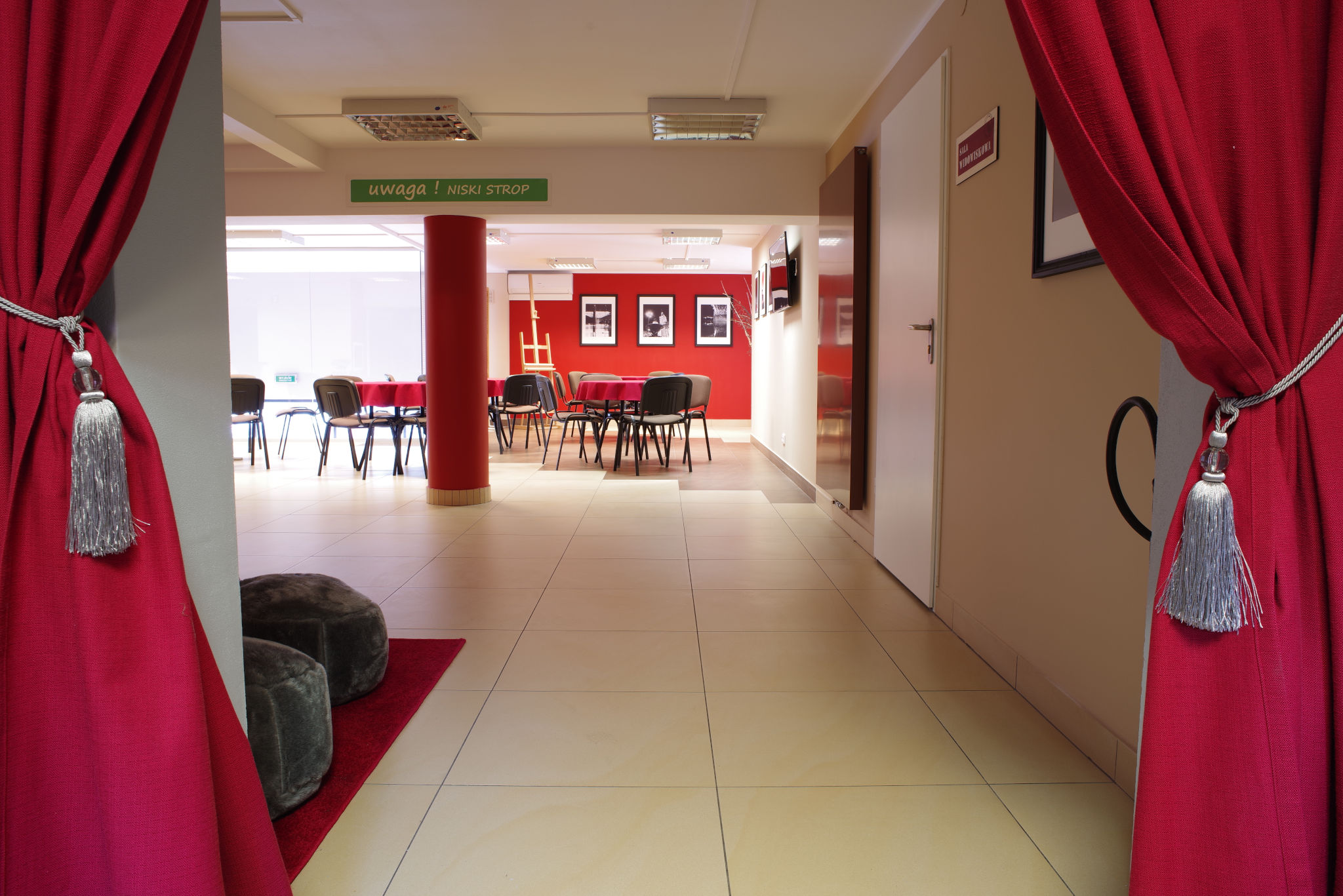 Sala na spotkanie. Widoczna kremowa podłoga, czerwona kolumna, rozstawione krzesła i stoły.  Widać czerwone kotary od strony wejścia na salę.