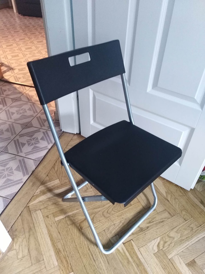 Plastikowe krzesło, rozłożone, czarne. Ustawione na drewnianej podłodze, w tle białe drzwi. 