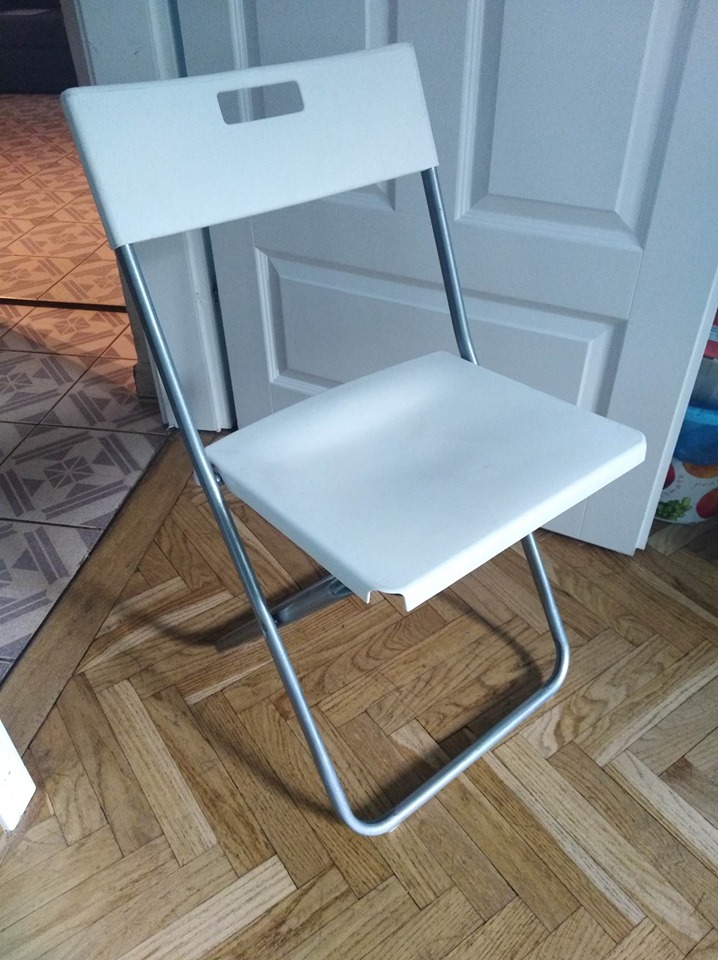 Plastikowe krzesło, rozłożone, białe. Ustawione na drewnianej podłodze, w tle białe drzwi. 