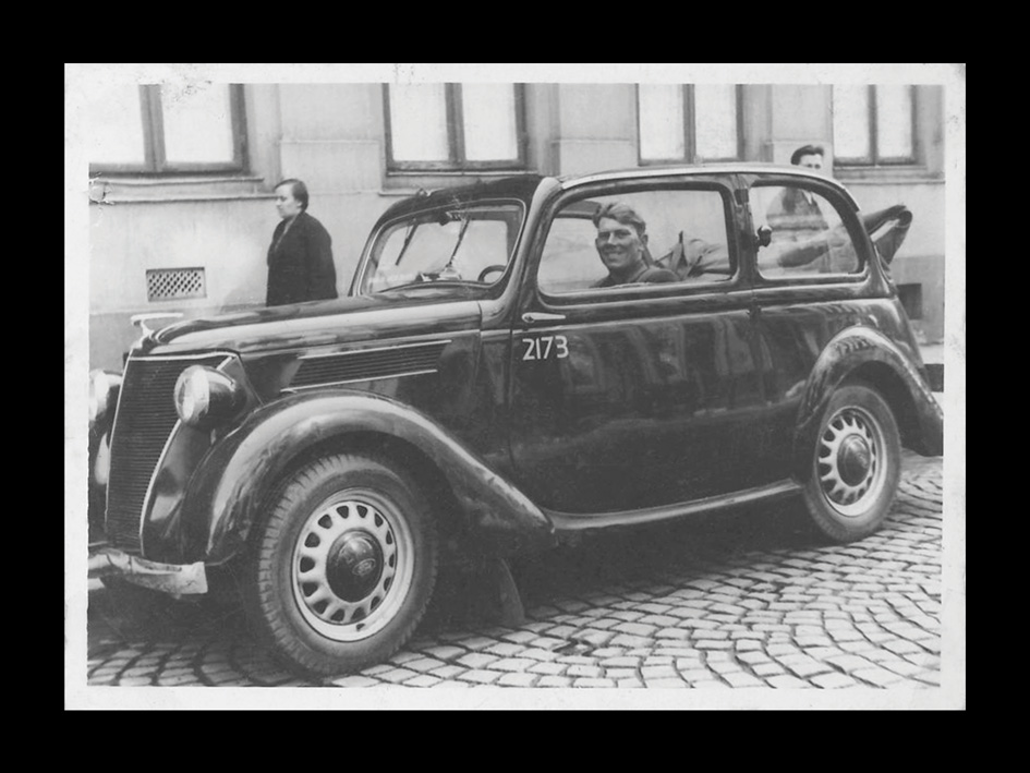 Zdjęcie czarnobiałe przedstawiające stary samochód stojący na ulicy. W środku auta znajduje się kierowca. 